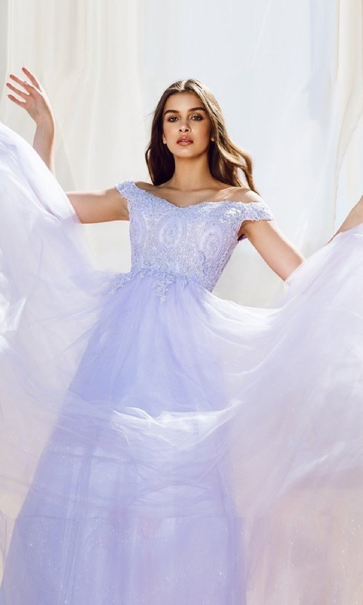 A woman in the Off-Shoulder Glitter Prom Ball Gown: Eureka 9899 stands before a light, neutral backdrop, lifting the skirt of her lavender dress adorned with lace embellishments.